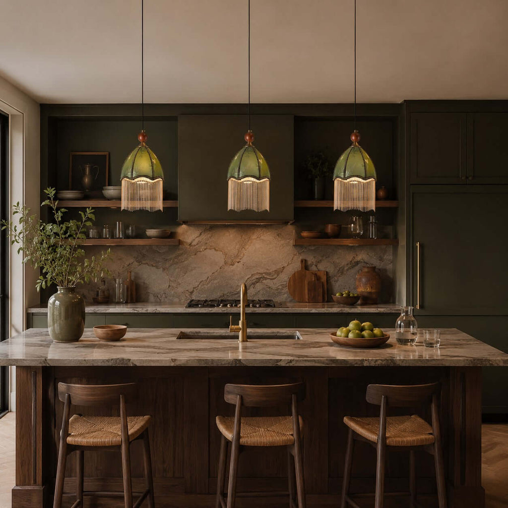 Three small Clérachic pendants with green shades and fringe hang over a stone island in a dark, moody kitchen.