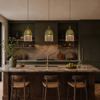 Three small Clérachic pendants with green shades and fringe hang over a stone island in a dark, moody kitchen.