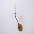 The Clérachic Eris wall lamp mounted and lit on a lavender-grey wall, its brass arm glowing warm.