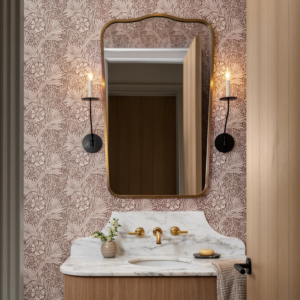 Clérachic's Eris wall lamp flanks a mirror in a dark finish above a marble vanity and floral wallpaper.