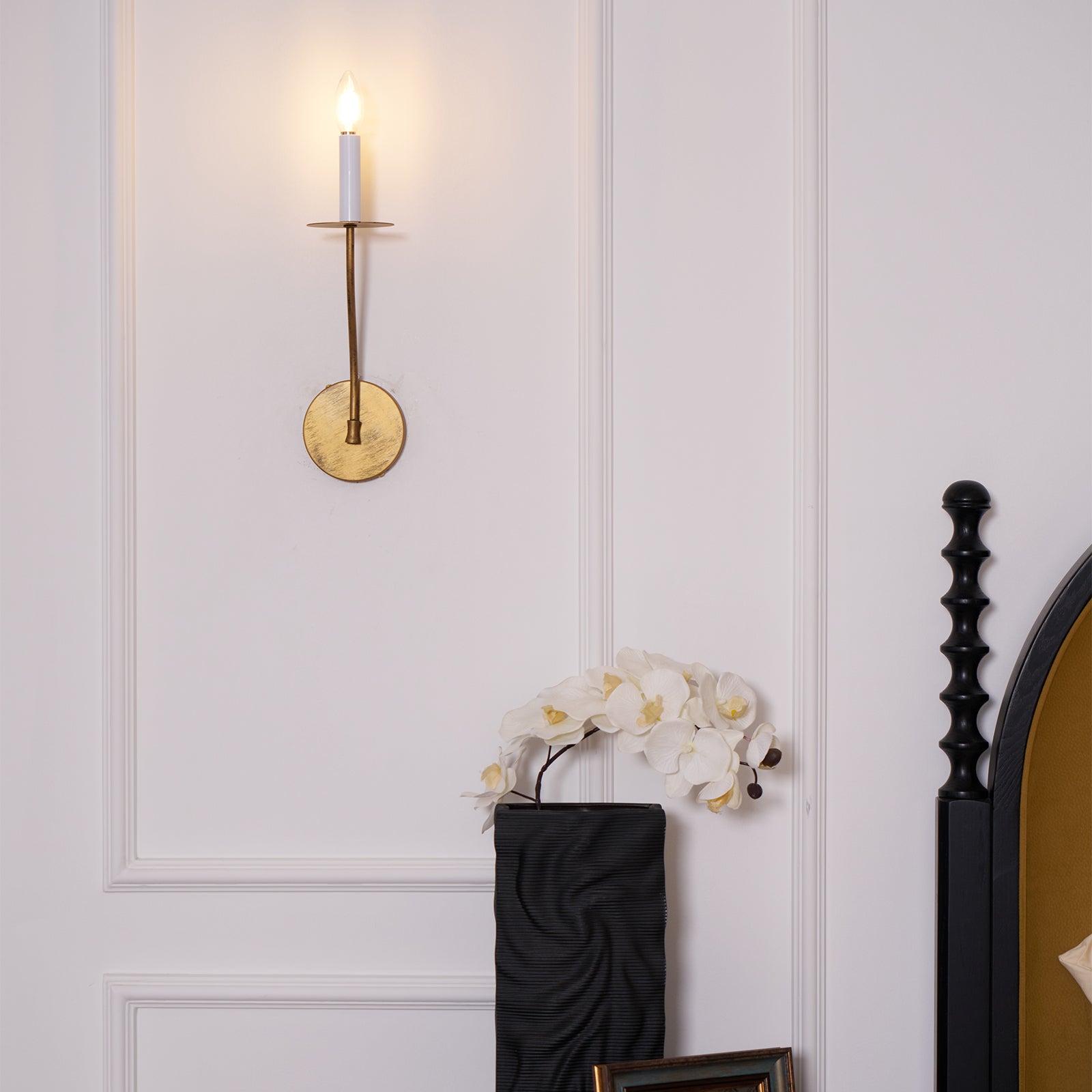 The Clérachic Eris wall lamp glows brass on a pink paneled bedroom wall beside an orchid on the nightstand.