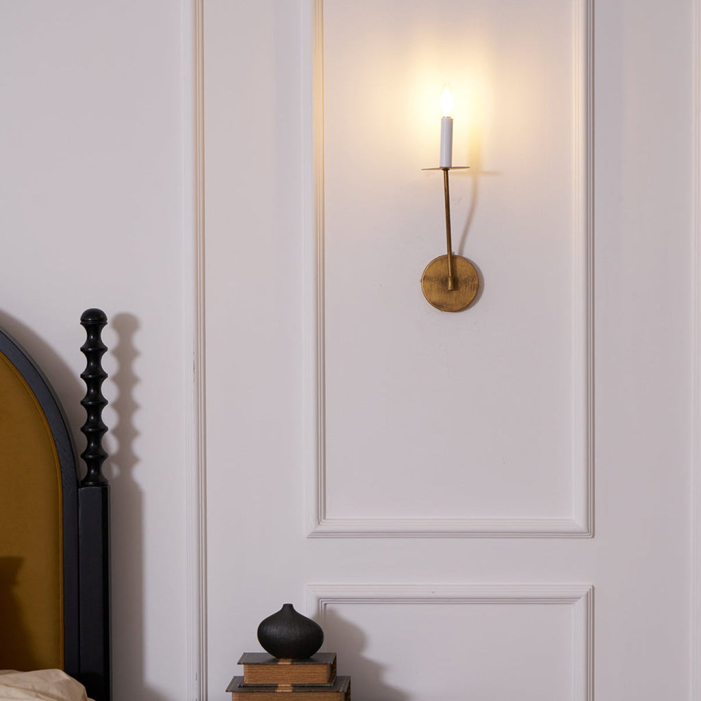 The Clérachic Eris wall lamp glows brass beside a black headboard, books stacked on the nightstand below.