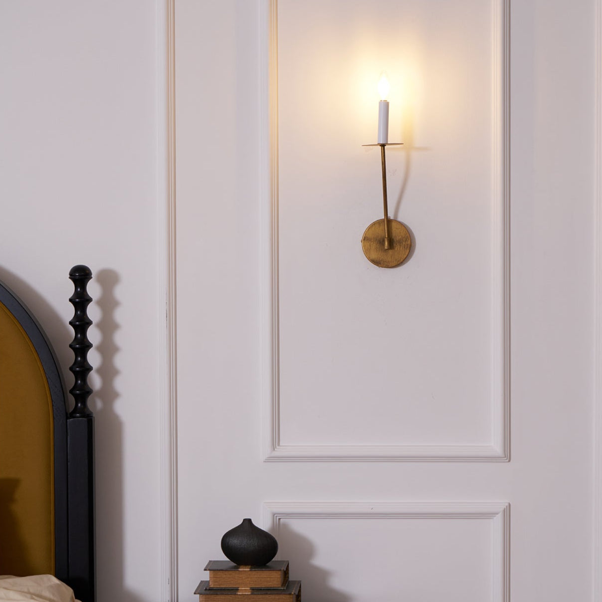 The Clérachic Eris wall lamp glows brass beside a black headboard, books stacked on the nightstand below.