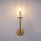 The Clérachic Eris wall lamp glows brass in close-up on a lavender-grey wall, its warm light prominent.