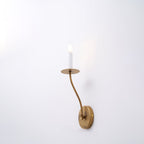 Clérachic's Eris wall lamp glows brass on a white wall, photographed from a wider angle.