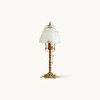 Table lamp with an antique brass stem, openwork base and a scalloped frosted glass shade, on a plain white backdrop, from Clérachic.