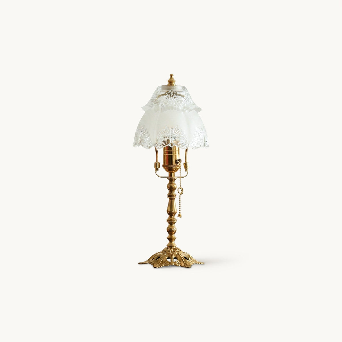 Table lamp with an antique brass stem, openwork base and a scalloped frosted glass shade, on a plain white backdrop, from Clérachic.