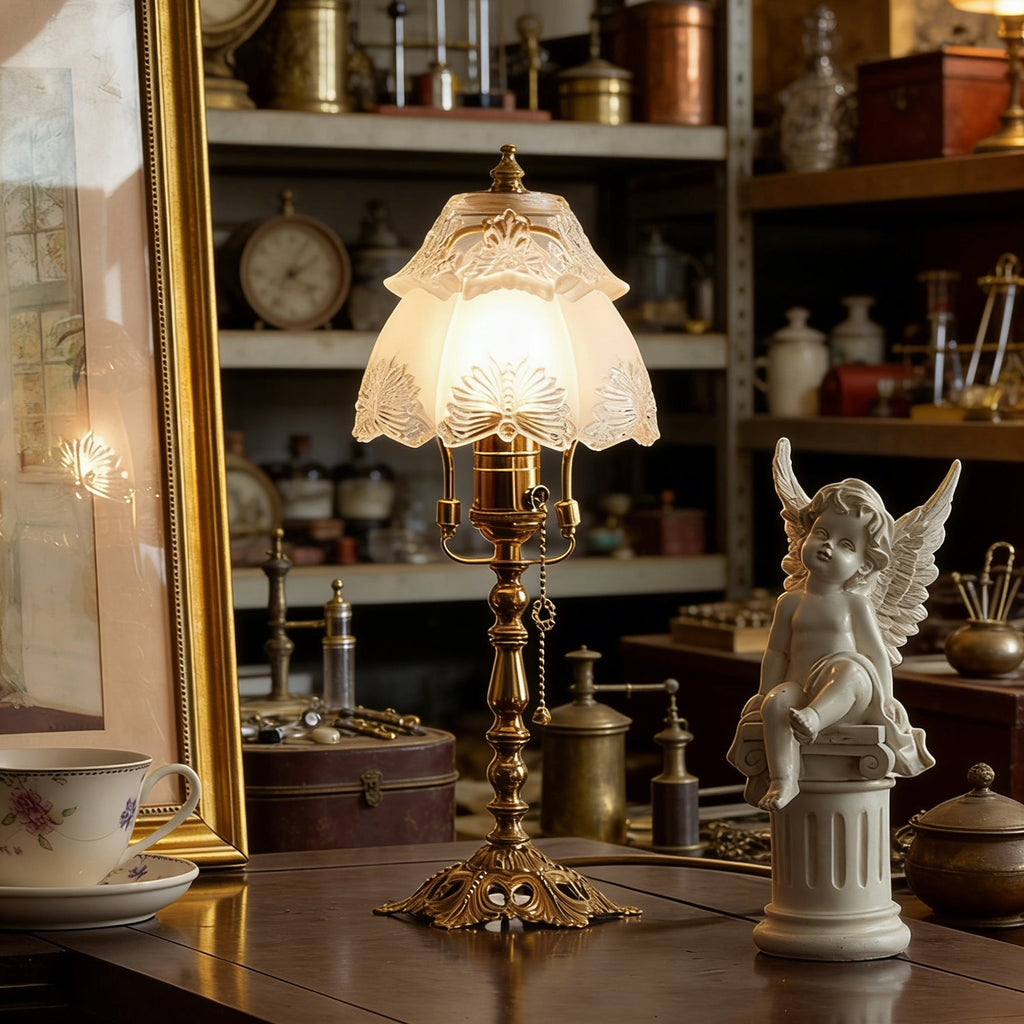 Lit table lamp with an etched scalloped shade on a dark wooden table, with a cherub figure, teacup and a shelf of curios behind, from Clérachic.