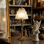 Lit table lamp with an etched scalloped shade on a dark wooden table, with a cherub figure, teacup and a shelf of curios behind, from Clérachic.