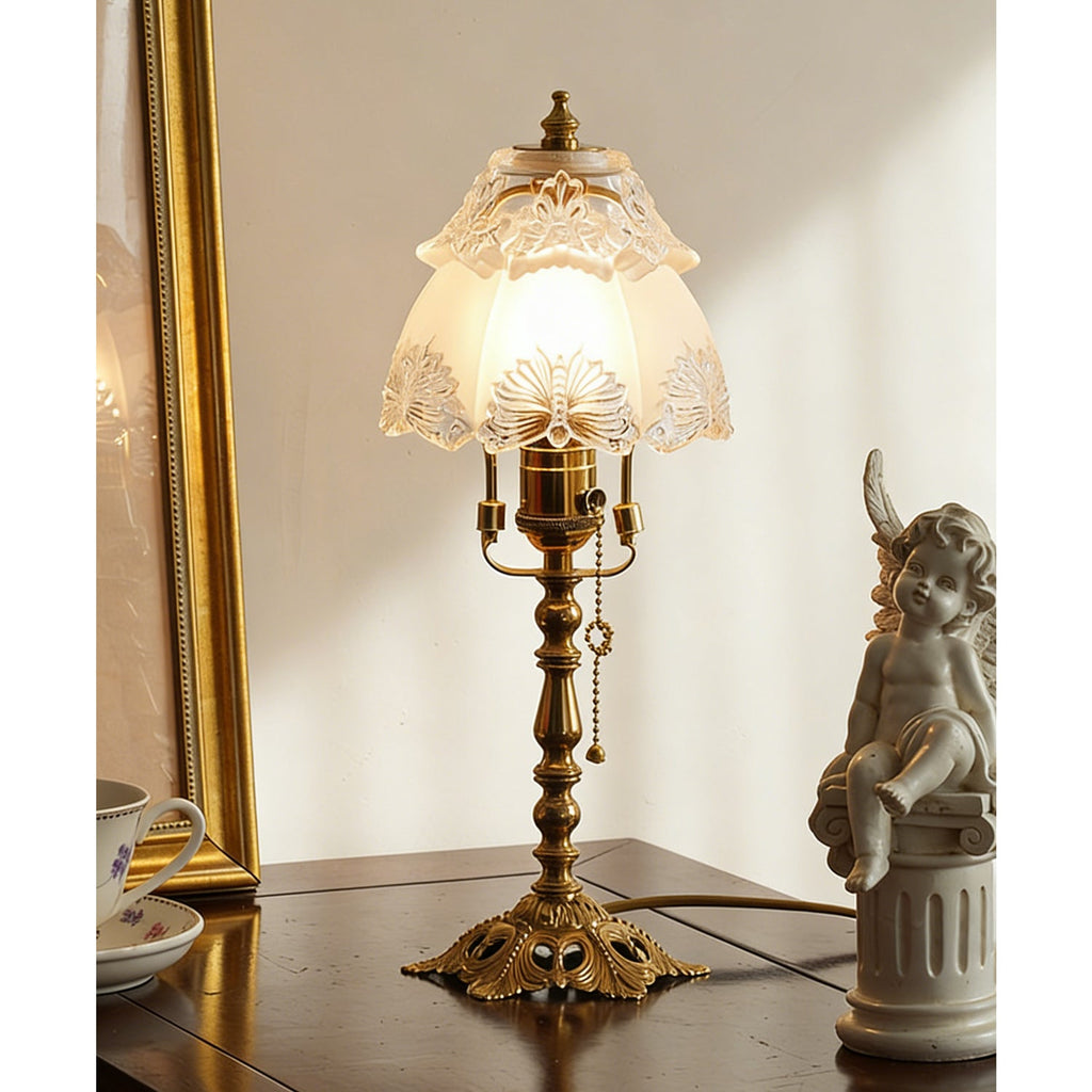 Glowing frosted glass shade with etched floral edging above a brass stem and pull chain, beside a gold frame and cherub, a Clérachic table lamp.