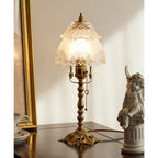 Glowing frosted glass shade with etched floral edging above a brass stem and pull chain, beside a gold frame and cherub, a Clérachic table lamp.