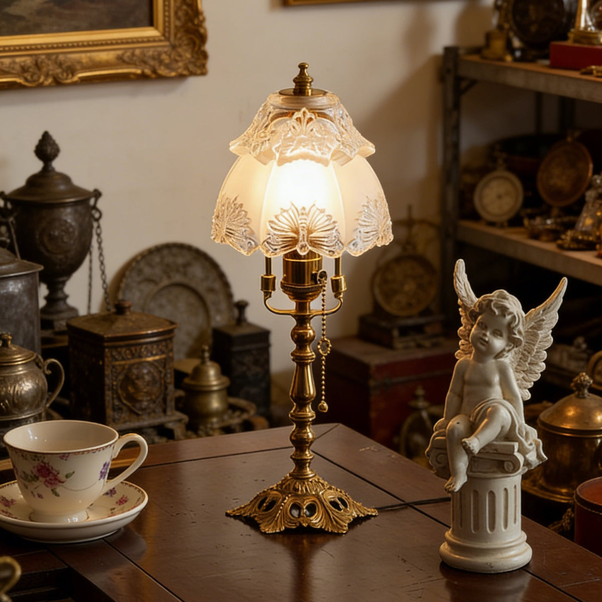 Warm-lit table lamp in a cluttered antique corner, with a floral teacup, brass ornaments and a white cherub figure, from Clérachic.