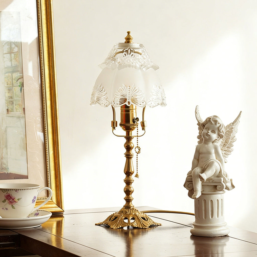 Unlit white scalloped glass shade on a slim brass stem, set on a polished table beside a cherub figure and gold frame, a Clérachic table lamp.