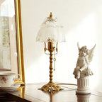 Unlit white scalloped glass shade on a slim brass stem, set on a polished table beside a cherub figure and gold frame, a Clérachic table lamp.