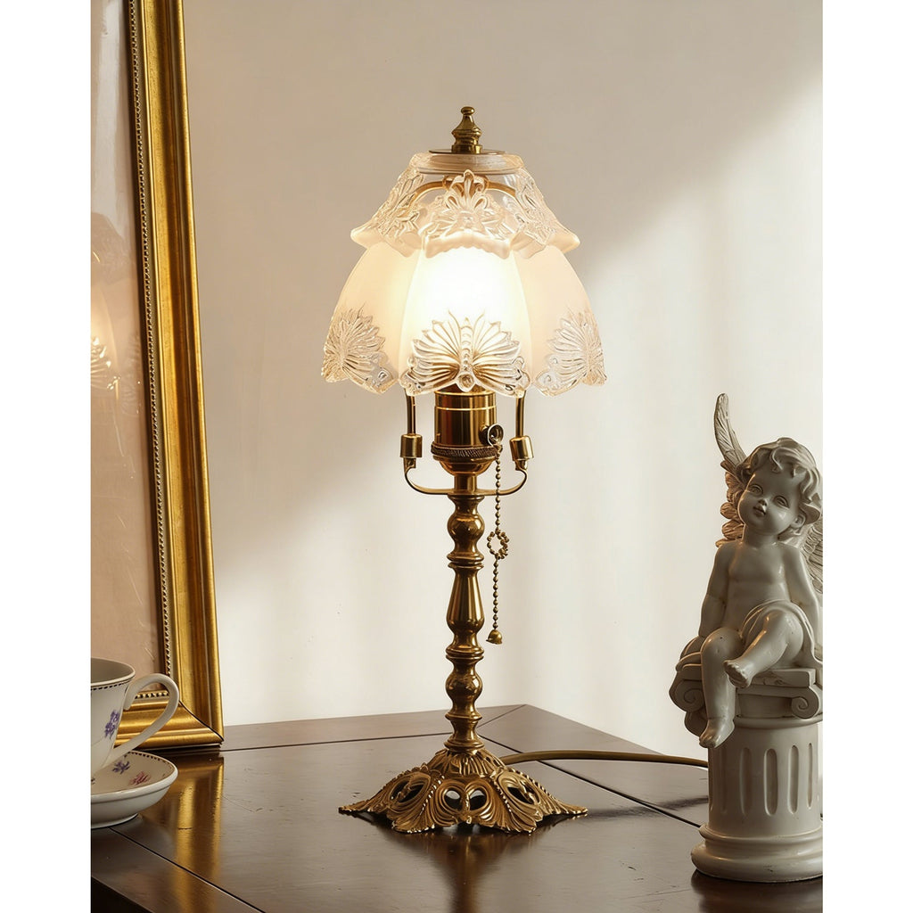 Table lamp lit warm against a cream wall, its brass base with pierced detail sitting on a dark table beside a cherub, from Clérachic.