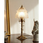 Table lamp lit warm against a cream wall, its brass base with pierced detail sitting on a dark table beside a cherub, from Clérachic.