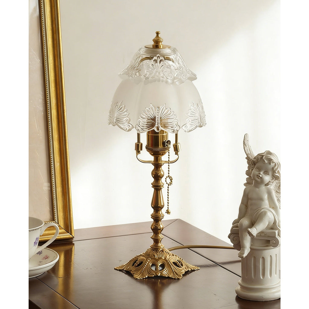 Slightly angled view of the brass lamp and frosted shade on a dark table, with a gold frame and a small cherub statue, a Clérachic table lamp.