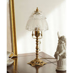 Slightly angled view of the brass lamp and frosted shade on a dark table, with a gold frame and a small cherub statue, a Clérachic table lamp.