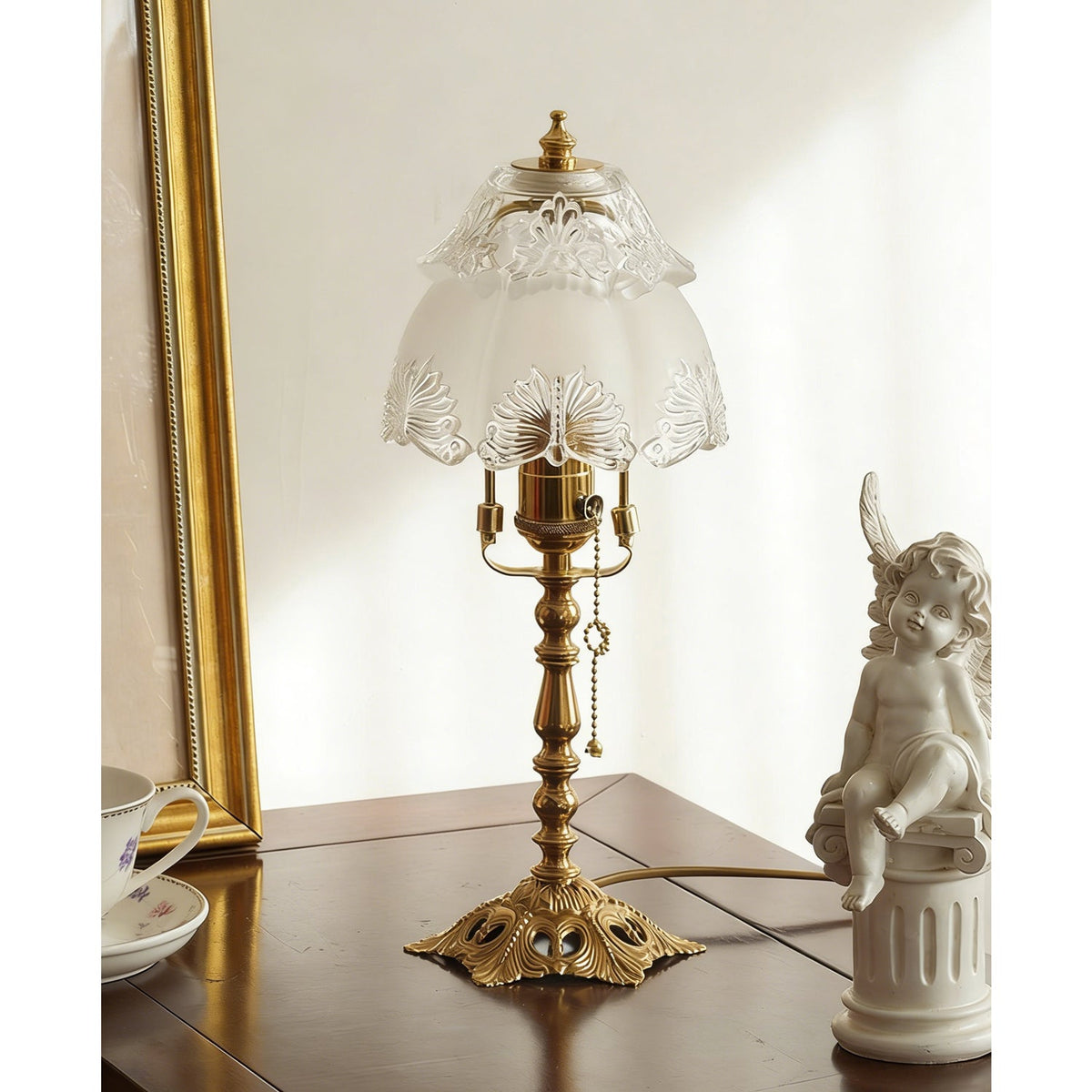 Slightly angled view of the brass lamp and frosted shade on a dark table, with a gold frame and a small cherub statue, a Clérachic table lamp.