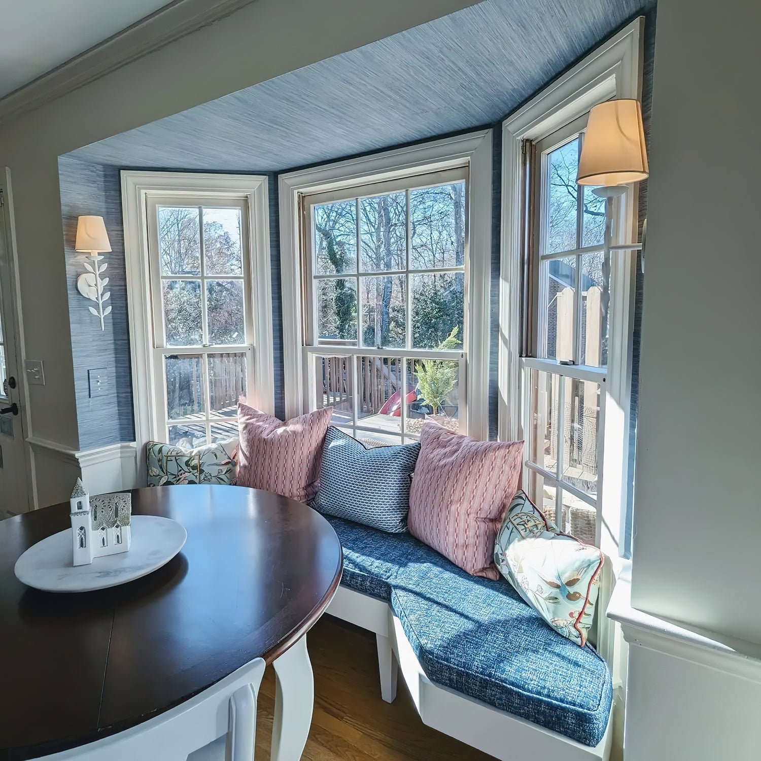 A Clérachic Evoque sconce mounted beside a bay window seat, its white shade glowing warmly at dusk.
