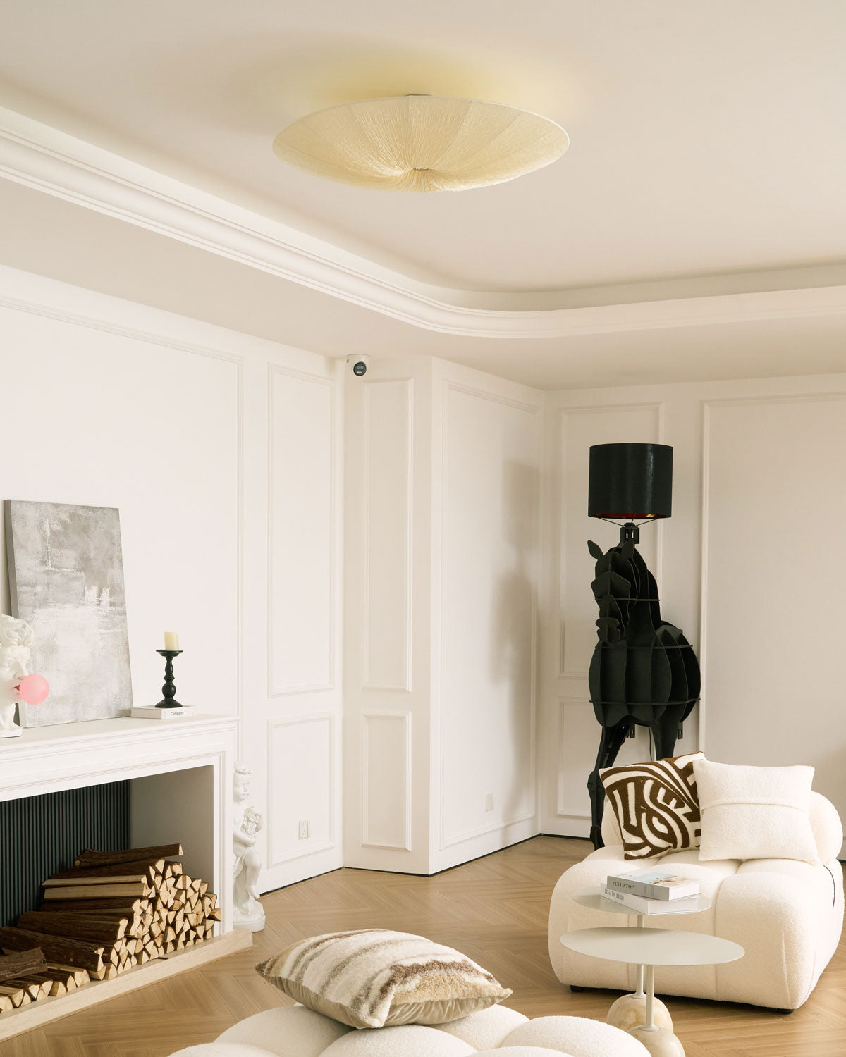 Unlit cream Clérachic fabric ceiling light under a coffered white ceiling, with a black horse sculpture lamp and a cream armchair below.
