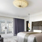 Clérachic pleated ceiling light overhead in a large bedroom, above a tufted dark headboard and a bed with a grey runner.