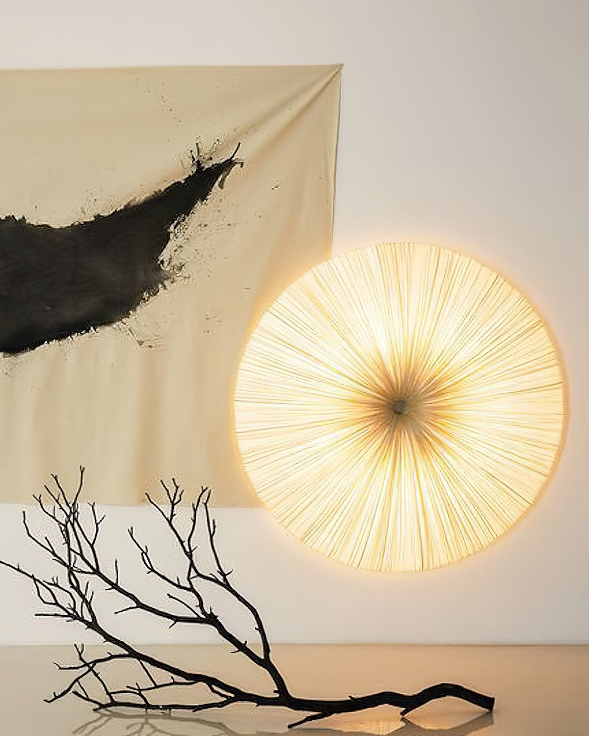 A round pleated Clérachic light fixed to a wall like a sun, glowing beside an ink-brushed hanging and a bare black branch.