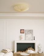 Clérachic pleated fabric ceiling light unlit, a pale disc above a white room with a fireplace, books and a small side table.