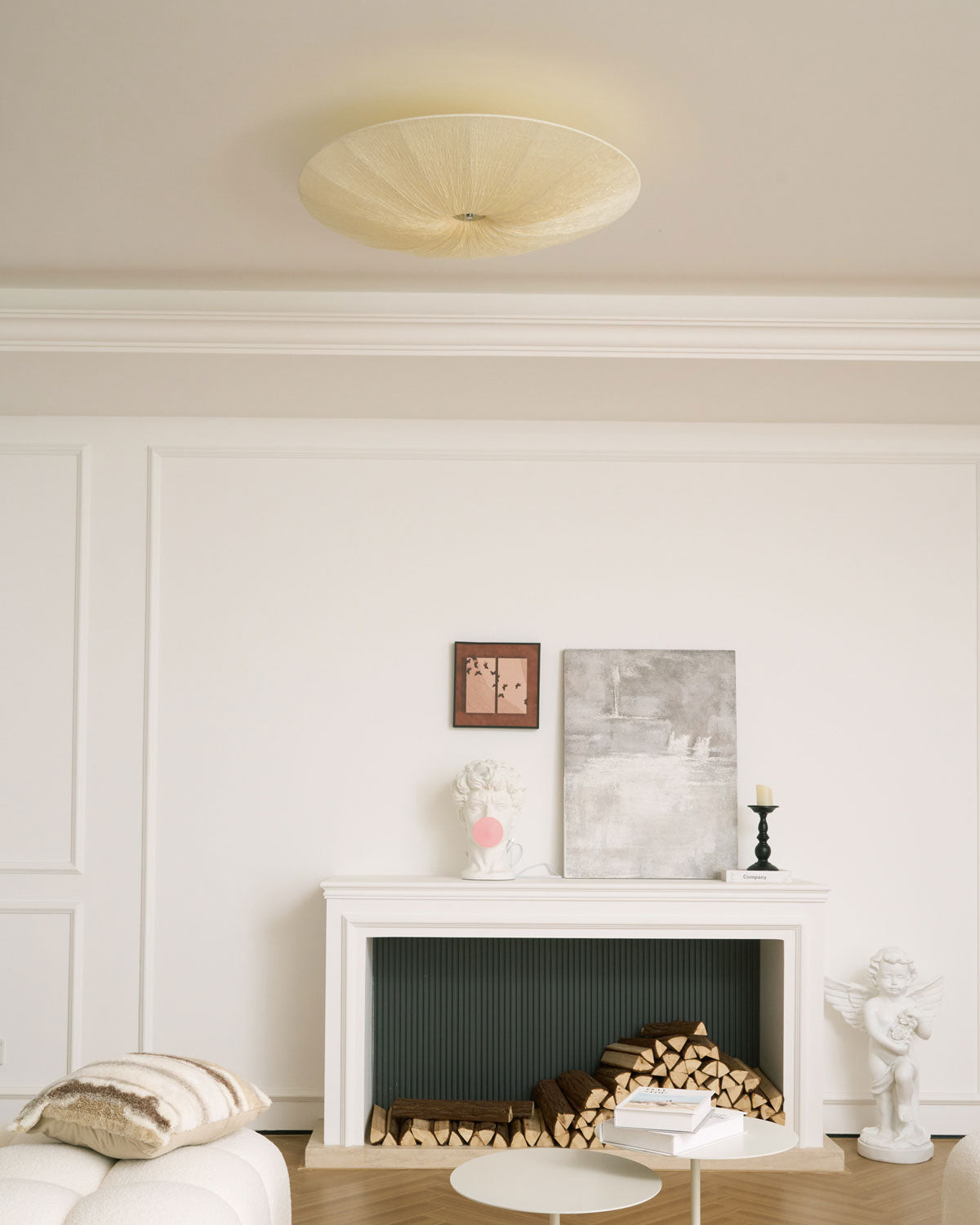 Clérachic pleated fabric ceiling light unlit, a pale disc above a white room with a fireplace, books and a small side table.