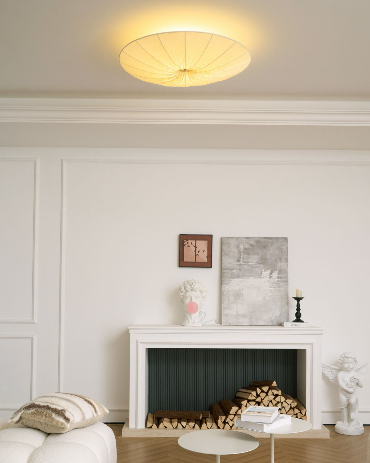 Clérachic pleated ceiling light lit with a soft halo on the ceiling, above a white mantel with logs and framed artwork.