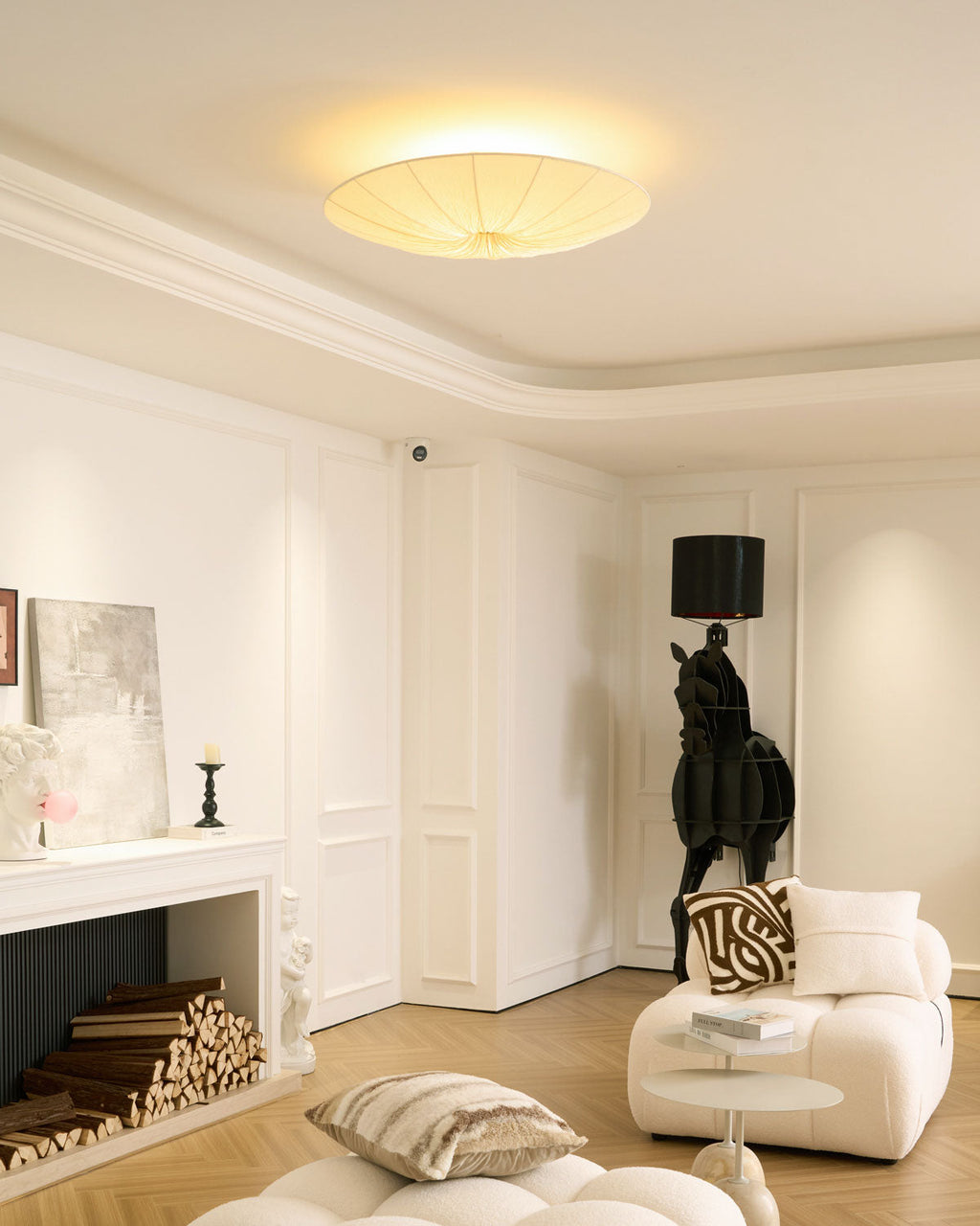 Clérachic fabric ceiling light glowing in a corner of a white room, with a stacked-log fireplace and a cream chair below.
