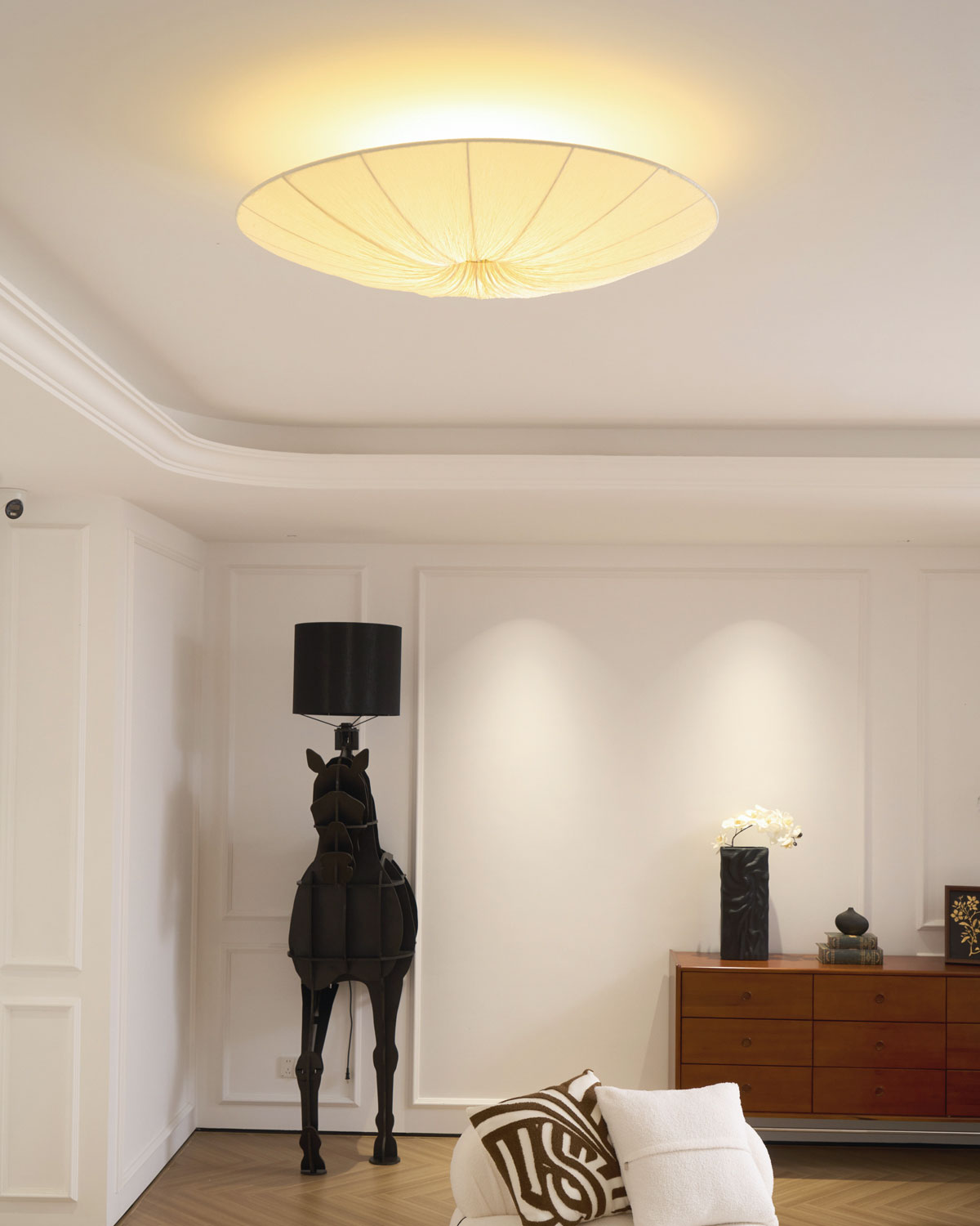 Clérachic fabric ceiling light lit above a panelled white room, a black horse lamp and a low wooden sideboard below.