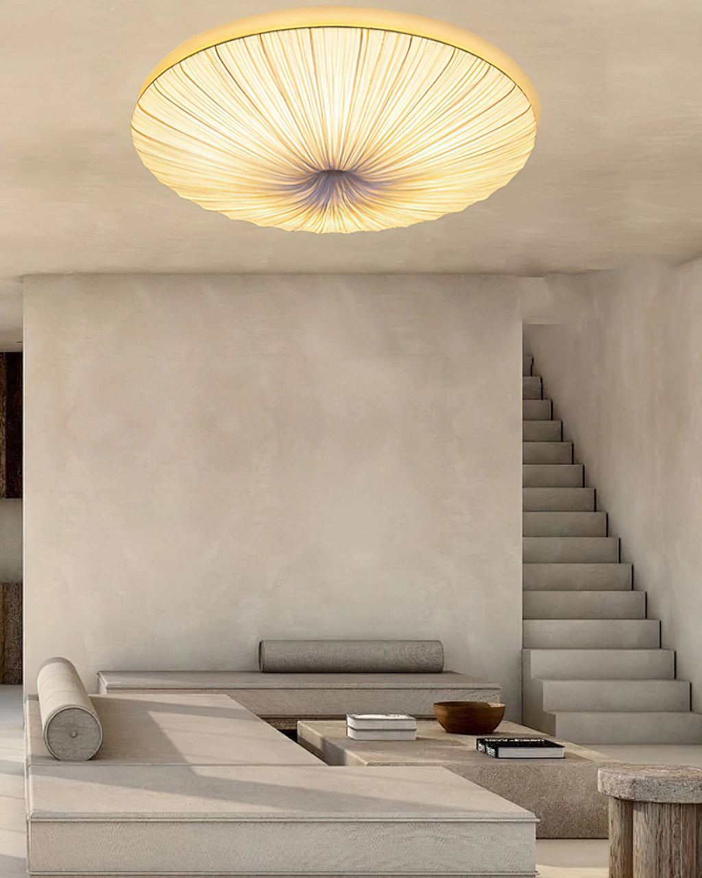 Lit Clérachic pleated ceiling light above a plaster room, with an open staircase and a long low bench in the background.