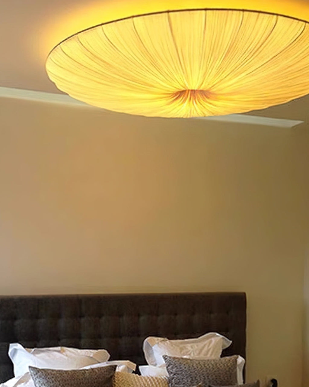 Looking up at a lit Clérachic fabric ceiling light above a warm beige wall, with a grey headboard and white pillows below.