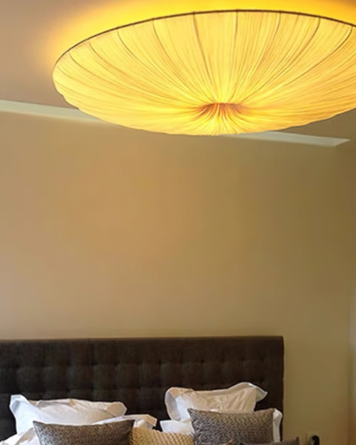 Looking up at a lit Clérachic fabric ceiling light above a warm beige wall, with a grey headboard and white pillows below.