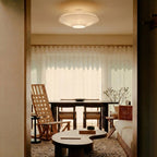 Lit pleated fabric ceiling light from Clérachic seen through a doorway, above a dining table and a low coffee table, with sheer curtains behind.