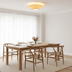 A Clérachic pleated ceiling light casting warm light over a pale room with a long oak dining table and woven-seat chairs.