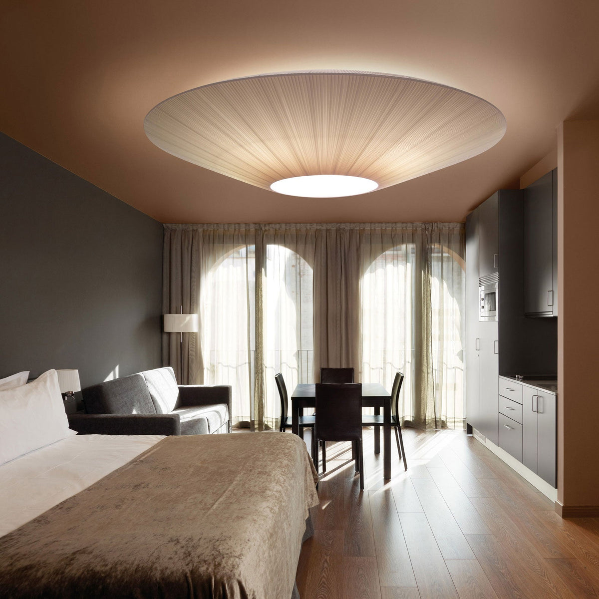 Large Clérachic pleated fabric ceiling light over a hotel-style bedroom, with a dark sofa, a kitchenette and arched windows behind.