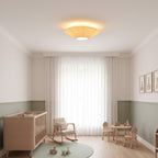 A Clérachic pleated ceiling light lit above a nursery with a wooden cot, a rocking chair, a small table and sheer curtains.