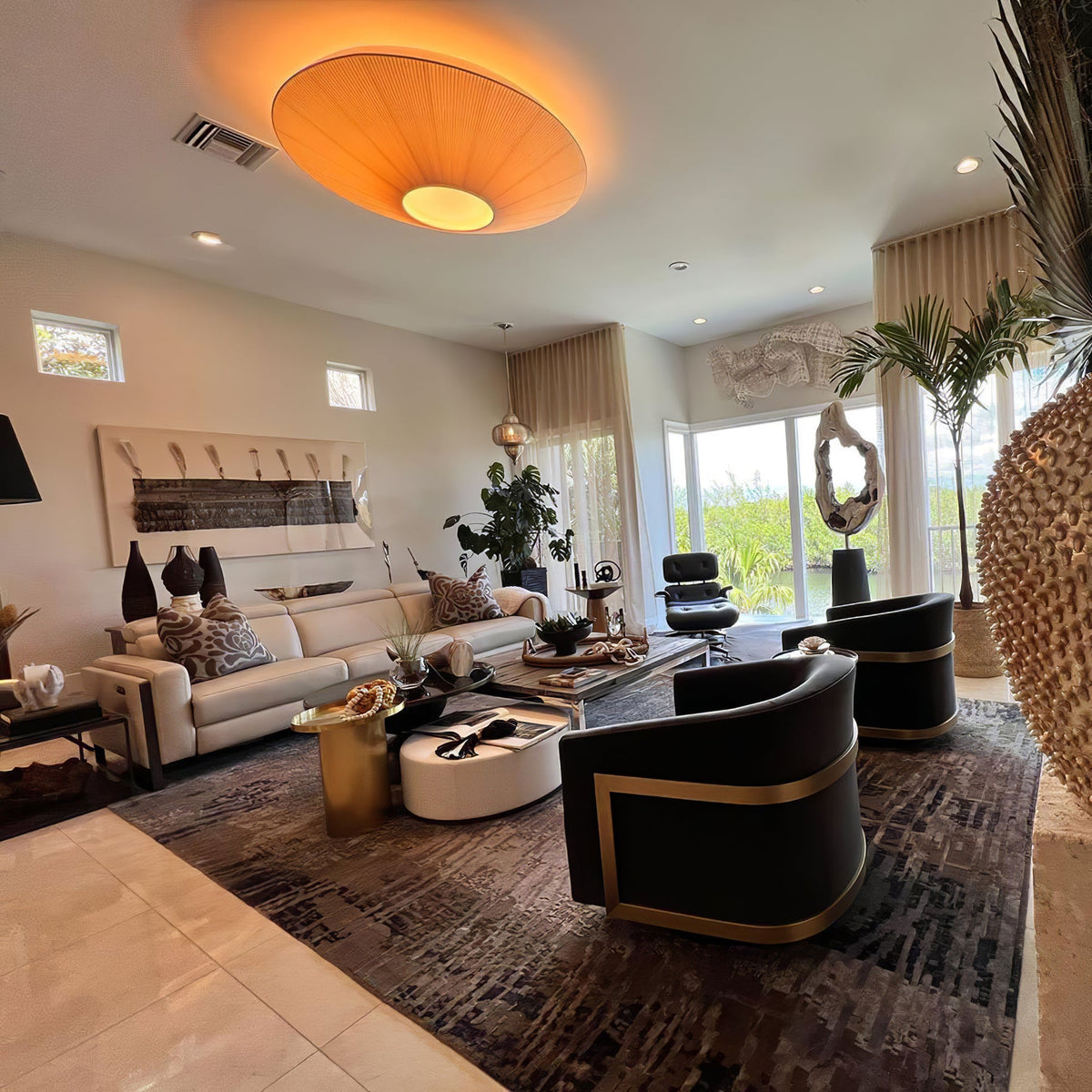 Orange-glowing Clérachic pleated ceiling light in a large living room with a beige sofa, black armchairs and a patterned rug.