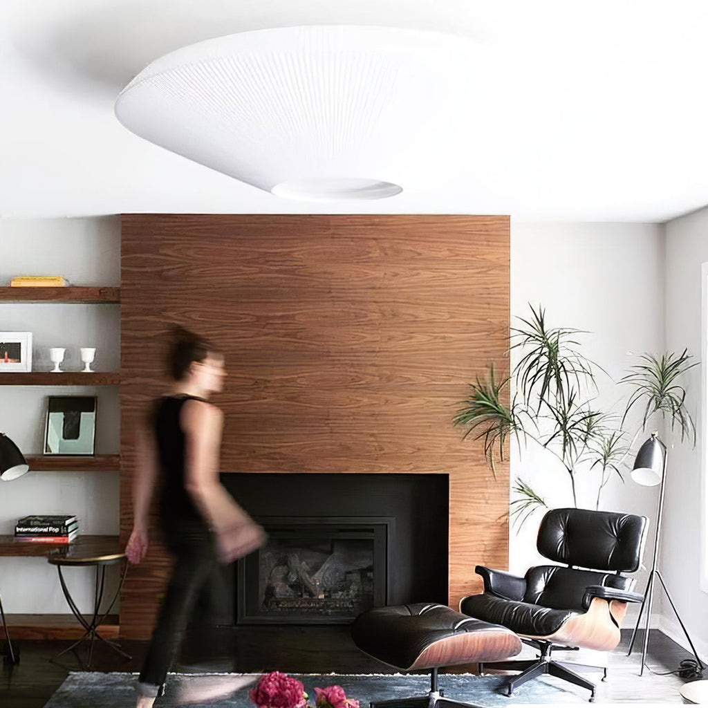 Underside of a white Clérachic pleated ceiling light above a walnut fireplace wall, with a leather lounge chair and a person walking past.
