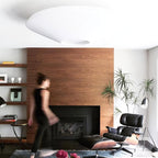 Underside of a white Clérachic pleated ceiling light above a walnut fireplace wall, with a leather lounge chair and a person walking past.