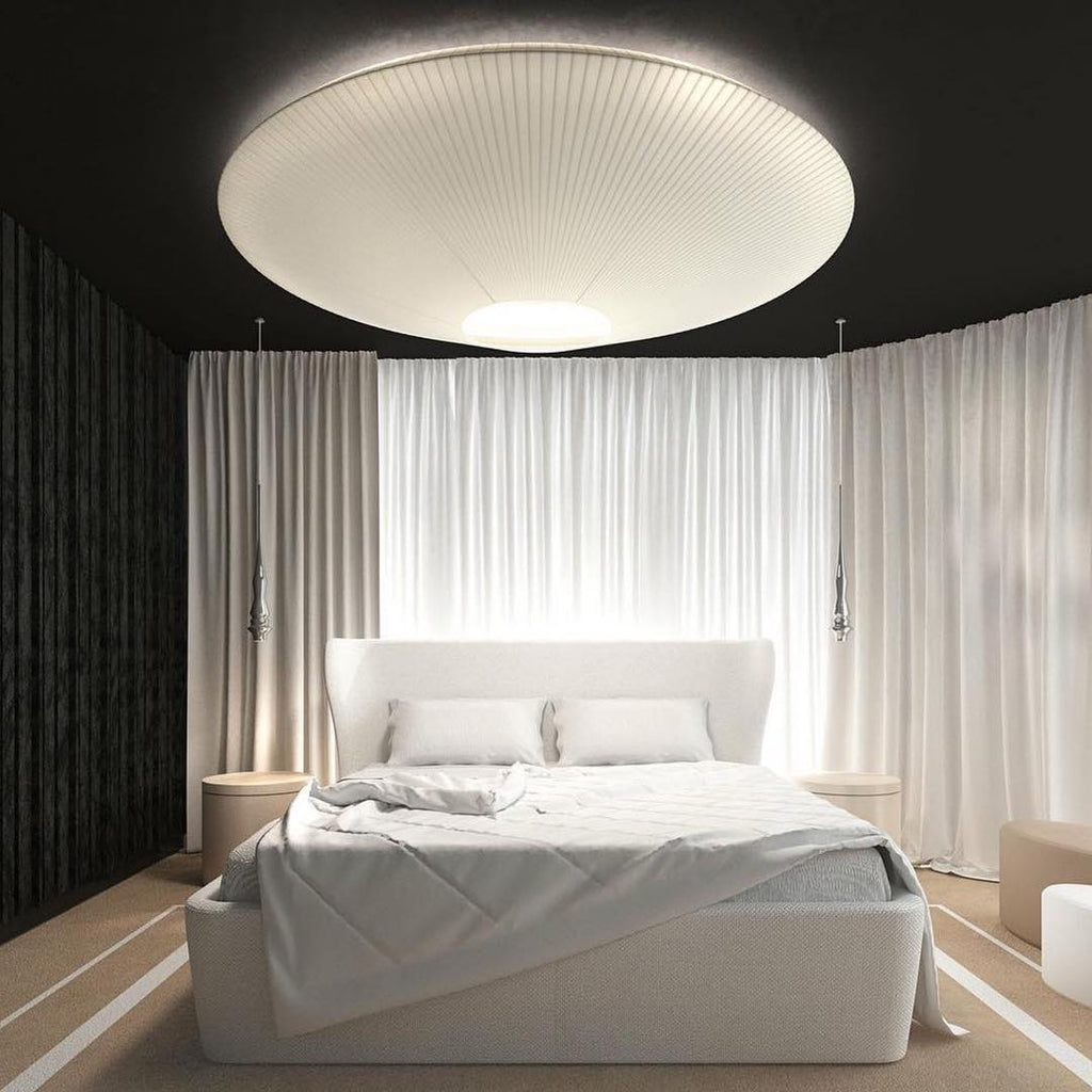 Big white Clérachic pleated ceiling light over a bed with white curtains drawn behind it, in a room with a dark ceiling.