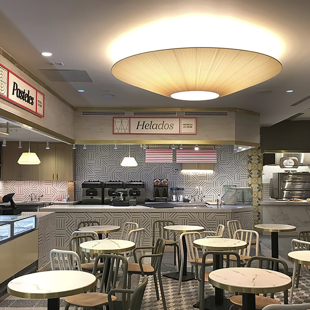 Large warm-toned Clérachic pleated ceiling light glowing above an ice-cream cafe, with round tables and metal chairs below.
