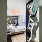 A Clérachic pleated ceiling light with a pinkish glow in a bedroom, seen past a tall sculptural mirrored panel.
