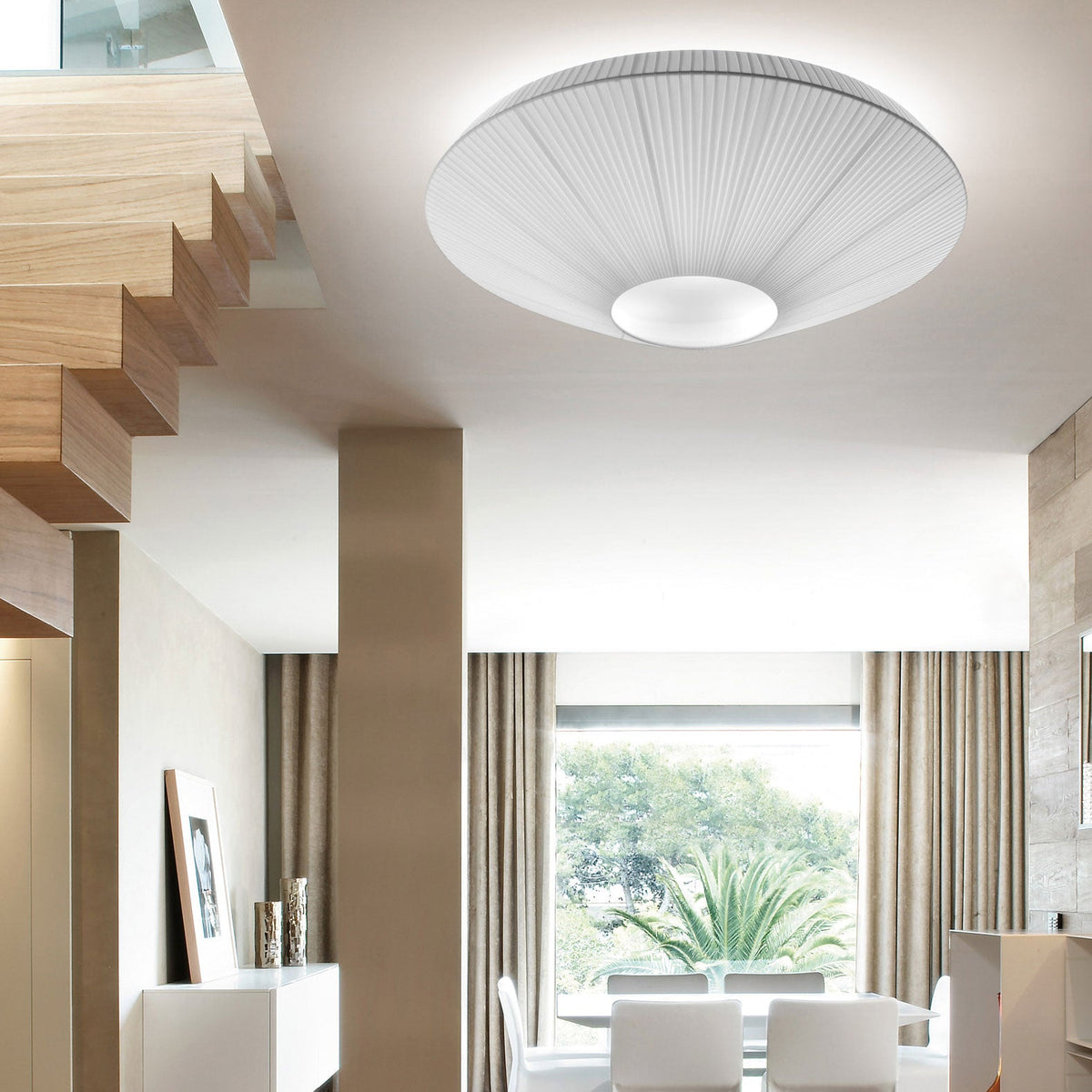 Close view from below of a white Clérachic pleated ceiling light with a round opal centre, above a stairwell and a dining area.