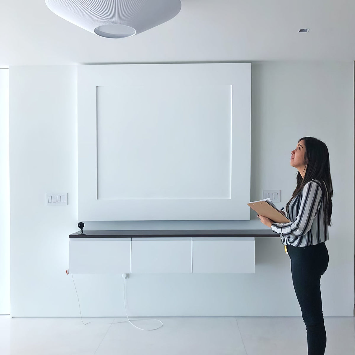 A woman looking up at a white Clérachic pleated ceiling light in a bright room with a built-in white wall unit.