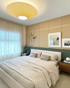 Yellow-toned Clérachic pleated ceiling light above a bedroom with timber wall panels, sheer curtains and framed pictures.