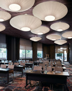 A cluster of Clérachic pleated ceiling lights in white at different sizes, hung from a dark ceiling above rows of tables in a conference room.
