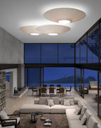 Three Clérachic pleated ceiling lights in ivory at different heights above a lounge with a cream sectional, a dining table and a night-time sea view.
